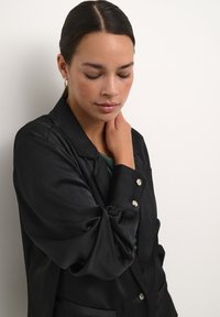 Blazer negru texturat cu nasturi argintii, având un guler clasic și două buzunare frontale, purtat peste un top verde.