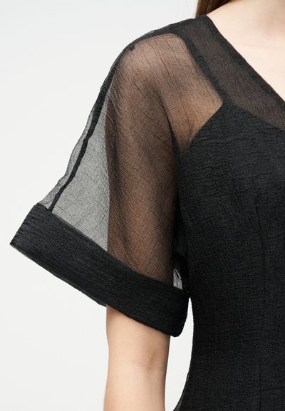 Μαύρο φόρεμα με υφή, διάφανο, κοντό με φουσκωτό μανίκι και φαρδύ μανσέτα, φορεμένο από ένα άτομο με ορατό μπράτσο και ώμο μπροστά σε απλό φόντο.