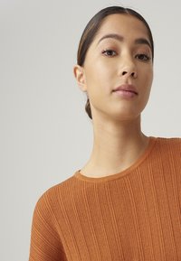 Ribbed, rust-colored sweater with a round neckline and soft texture, featuring long sleeves and a relaxed fit.