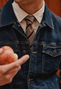 Denim jakna s bronastimi gumbi, prekrita s kremasto srajco, ki ima rjav prugast kravato. Ročna drži rdečo jabolko v ospredju.