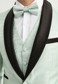 Veste de costume paisley vert menthe clair avec revers noirs, associée à un nœud papillon assorti et une chemise blanche en dessous.