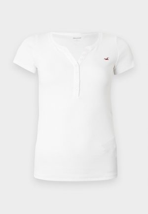 Balta īsroku henleja tipa krekls no ribotas auduma, ar pogu rindu un nelielu sarkanu logotipa akcentu krūtīs.