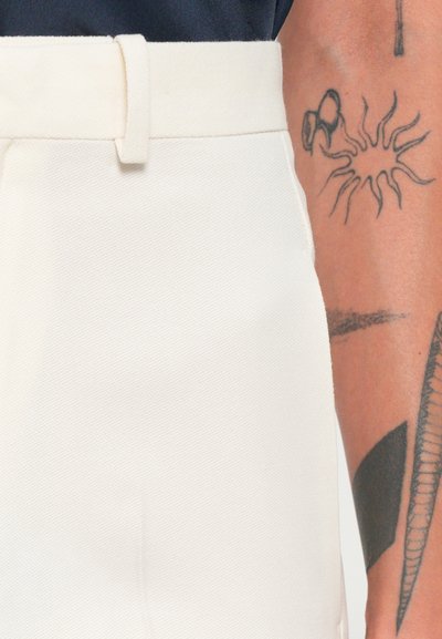 White cotton trousers with a straight cut, featuring a belt loop and a smooth texture. Visible arm with tattoos in the background.