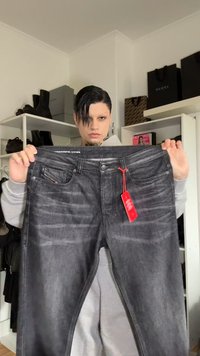 Jeans pretos com um acabamento ligeiramente desbotado, bolsos frontais e uma etiqueta vermelha presa. A cintura apresenta o nome da marca em letras brancas.