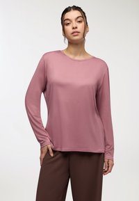 Długi rękaw w stonowanym różu, wykonany z gładkiej tkaniny. Posiada okrągły dekolt i luźny krój, zestawiony z brązowymi spodniami.