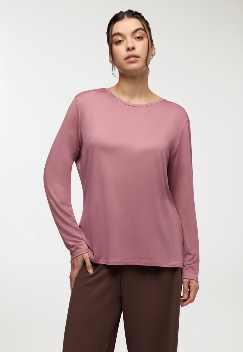 Długi rękaw w stonowanym różu, wykonany z gładkiej tkaniny. Posiada okrągły dekolt i luźny krój, zestawiony z brązowymi spodniami.