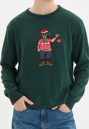 Dunkelgrüner Pullover mit einem Cartoon-Affen, der einen roten Weihnachtspullover und Hut trägt und ein rotes Ornament hält, getragen von einer Person mit Händen in den Taschen.