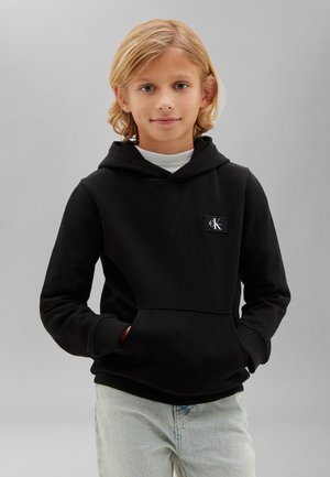 Blondt barn iført en sort Calvin Klein hættetrøje og lyseblå jeans står med hænderne i lommerne mod en ensfarvet grå baggrund.