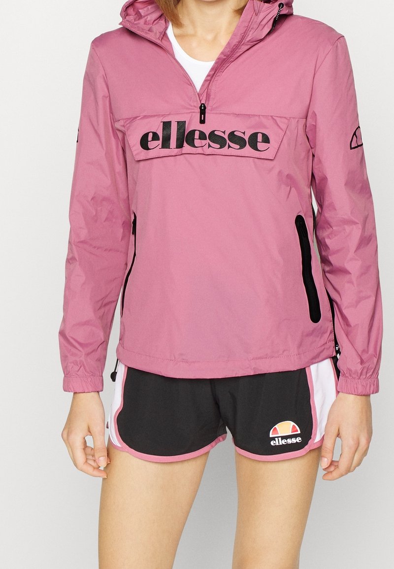 Rosa vindjacka med en framlappficka, elastiska ärmslut och svart logodetalj. Åtföljs av svarta shorts med rosa och vita accenter.