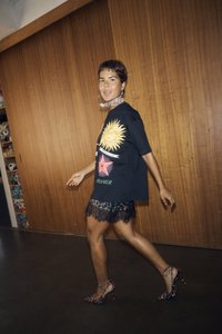 Tricou oversized negru cu un grafic al soarelui, asortat cu o fusta mini din dantelă neagră și tocuri înalte cu imprimeu de leopard. Ten uniform și păr scurt.