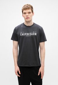 Must mustärgne lühikeste varrukatega puuvillane T-särk, millel on valge "CALVIN KLEIN" logo koos tekstiga "Asutatud 1968", ümar kaelus ja tavaline lõige.