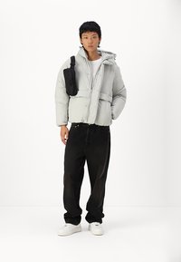 Chaqueta ligera de plumas gris claro con capucha, que presenta un cierre de cremallera y dobladillo elástico. Combinada con vaqueros baggy negros y zapatillas blancas.