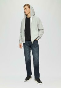 Haut à capuche gris zippé, t-shirt noir en dessous, jean bleu foncé et baskets noires avec semelles blanches, se tenant contre un fond clair.