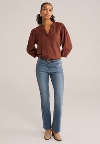 Blouse boutonnée couleur rouille avec des manches bouffantes et un col plissé, associée à un jean ajusté bleu clair et des bottines beige.