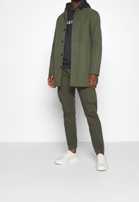Cappotto verde oliva con chiusura a bottoni, indossato sopra un felpa blu navy e pantaloni coordinati, abbinato a sneakers bianche. Tessuto liscio, vestibilità rilassata.