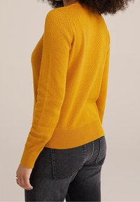 Mosterdgele trui met een textuursteekpatroon, slanke pasvorm en geribde zoom, gecombineerd met donkergrijze jeans.