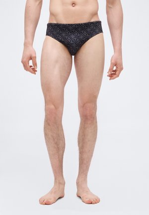 Male model wearing black briefs with a repeated white text pattern, standing barefoot against a plain white background.