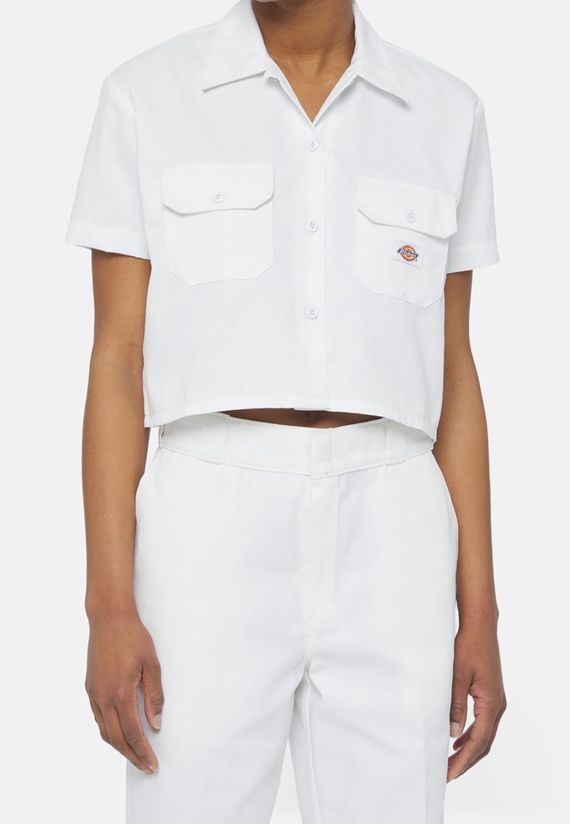 Personne portant une chemise blanche à manches courtes raccourcie avec deux poches sur la poitrine et un pantalon blanc taille haute assorti.