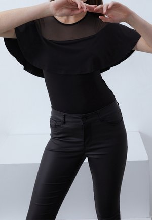 Femme portant un haut noir ajusté avec empiècement transparent et large détail à volants, assorti à un pantalon noir taille haute, posant les bras levés.