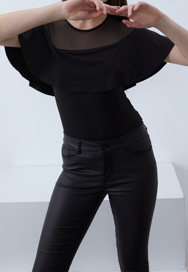 Femme portant un haut noir ajusté avec empiècement transparent et large détail à volants, assorti à un pantalon noir taille haute, posant les bras levés.