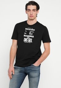 Čierne bavlnené tričko s bielym grafickým textom, okrúhlym výstrihom a krátkymi rukávmi. Model stojí uvoľnene v kombinácii s modrými džínsami.