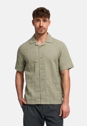 Giovane uomo che indossa una camicia a maniche corte color oliva chiaro con bottone e pantaloni grigio scuro, in piedi con una mano in tasca.