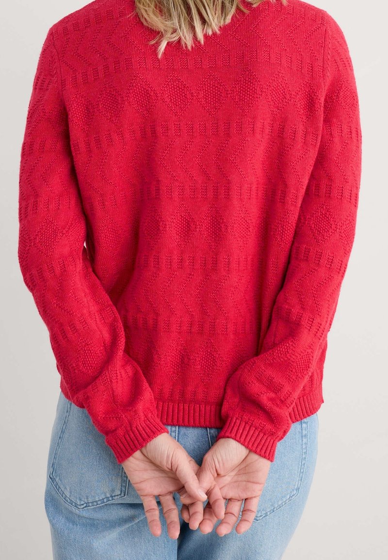 Personne aux cheveux blonds se tenant de dos, portant un pull rouge texturé et un jean bleu clair, les mains jointes dans le dos.