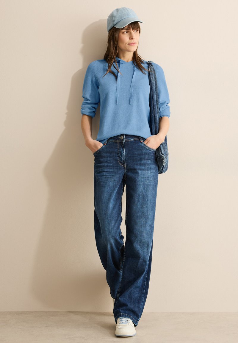 Felpa con cappuccio azzurro chiaro e lacci, abbinata a jeans blu scuro a vestibilità ampia e sneakers bianche, accessoriata con un cappellino azzurro chiaro.