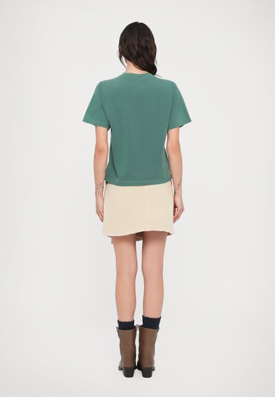 Πράσινη κοντομάνικη μπλούζα, σε άνετη γραμμή, συνδυασμένη με μπεζ φούστα. Καφέ μποτάκια στους αστραγάλους με μπλε πλεκτές λεπτομέρειες ολοκληρώνουν την εμφάνιση.