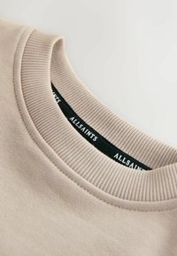 Beige sweatshirt met een geribbelde ronde hals en een zwart label waarop "ALLSAINTS" in witte tekst staat. Gladde katoenen stof met minimale stiksels.