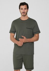 Camisa de manga corta verde oliva con un bolsillo en el pecho y ribete negro, combinada con pantalones cortos a juego que presentan acentos negros. Textura de tela suave.