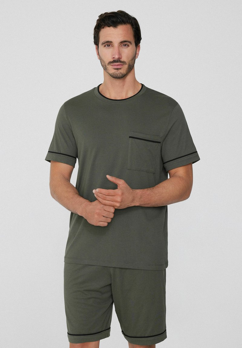 Camisa de manga corta verde oliva con un bolsillo en el pecho y ribete negro, combinada con pantalones cortos a juego que presentan acentos negros. Textura de tela suave.