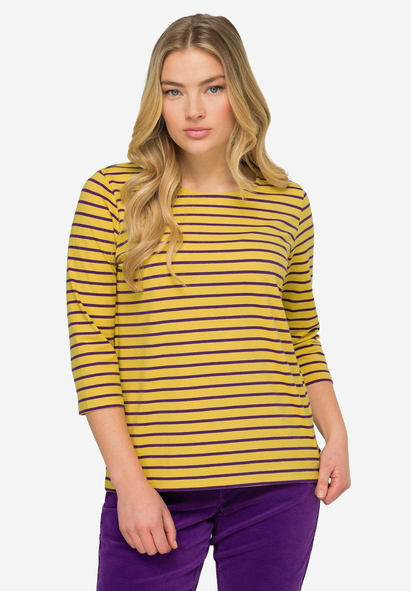 Gelbes Langarmshirt mit lila horizontalen Streifen; runder Halsauschnitt; weicher Stoff; kombiniert mit lila Kordhosen.