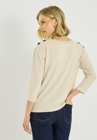 Top beige y ligero con mangas tres cuartos, con detalles de botones en el hombro. Combinado con jeans de mezclilla oscura.
