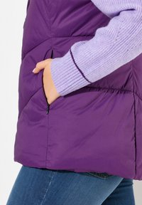 Lila Pufferweste mit glänzender Textur, ausgestattet mit einer Reißverschlusstasche und einem leicht abgerundeten Saum. Ribbed lavender Ärmeldetails sind sichtbar.