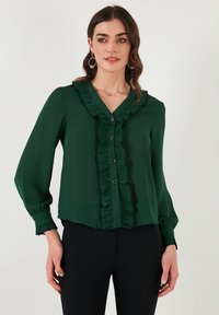 Grüne Bluse mit langen Ärmeln, Rüschendetails entlang des Ausschnitts und der Front, Knopfverschlüsse und geraffte Bündchen. Glatte Stofftextur.