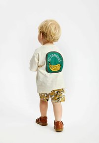 Chemise beige à manches courtes avec un graphique "Go Bananas" vert et rouge, associée à un short jaune avec des motifs de bananes. Sandales marron.