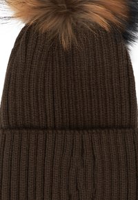 Beanie lavorato a maglia marrone con una texture a coste e un grande pompon soffice sulla parte superiore, caratterizzato da diverse tonalità di marrone sulla punta.
