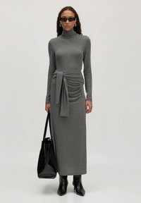 Grijs, geribbeld, lange mouwen jurk met een hoge hals en tailleband detail. Model houdt een zwarte tote bag vast en draagt strakke zwarte enkellaarsjes.