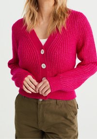 Ljusrosa stickad cardigan med djup V-ringning, rippad textur och tre stora vita knappar. Matchad med olivgröna byxor.