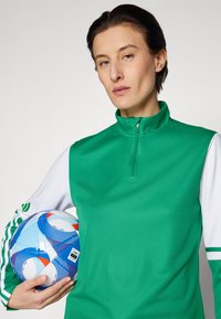 Grøn zip-neck atletisk top med hvide ærmer, som holder en multicolorer fodbold. Stoffet ser glat og tætsiddende ud, med subtile detaljer.