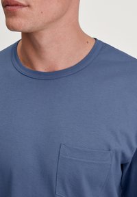Blauwe katoenen T-shirt met een ronde halslijn en een borstzak. De stof heeft een gladde textuur en subtiele stikdetails.