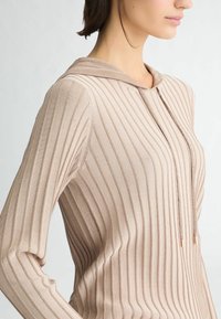 Haut beige à capuche avec une texture côtelée verticale, coupe ajustée et accents à cordon. Présente des manches longues et un tissu doux et lisse.