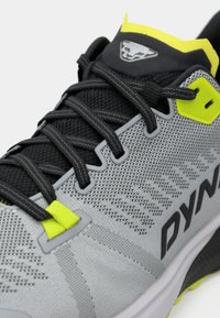 Γκρι αθλητικό παπούτσι με επάνω μέρος από διχτυωτό υλικό, μαύρες κορδόνες, νεον κίτρινες λεπτομέρειες και υφή σόλας με έντονο λογότυπο της μάρκας.