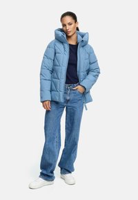 Ljusblå puffjacka med kviltat mönster, stor huva och dragkedja fram. Kombinerad med vida blå jeans och vita sneakers.