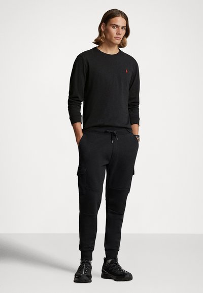 Svart longsleeve genser med brodert logo, kombinert med svarte cargo treningsbukser med lommer, båret med svarte treningssko.