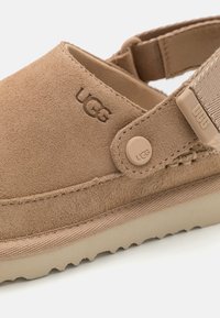 Suedefärgade UGG-sandaler med justerbar rem, märkknapp och texturerad sula på en vit bakgrund.