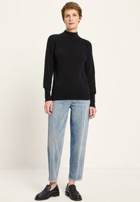 Person, der einen schwarzen Langarm-Turtleneck-Pullover, hellblaue gerade Jeans und schwarze LederSlipper trägt, steht vor einem hellen Hintergrund.