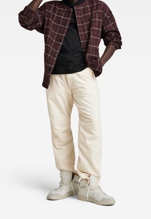 Man draagt een bordeauxrood geruit jasje over een zwart shirt, crèmekleurige broek en witte hoge sneakers, staat met één hand in de zak.