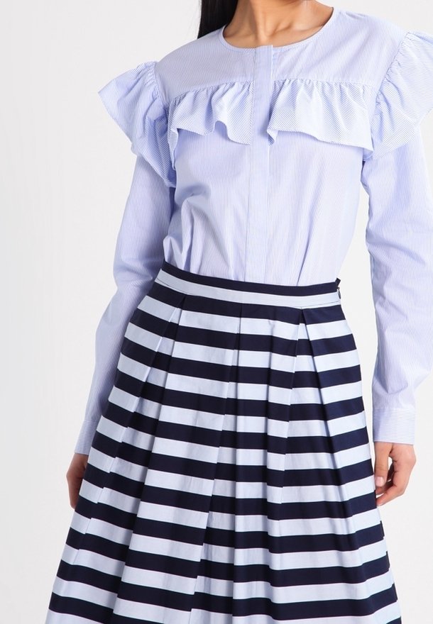 Femme portant un chemisier bleu clair à manches longues avec des volants et une jupe plissée à rayures horizontales bleu marine et blanches taille haute.